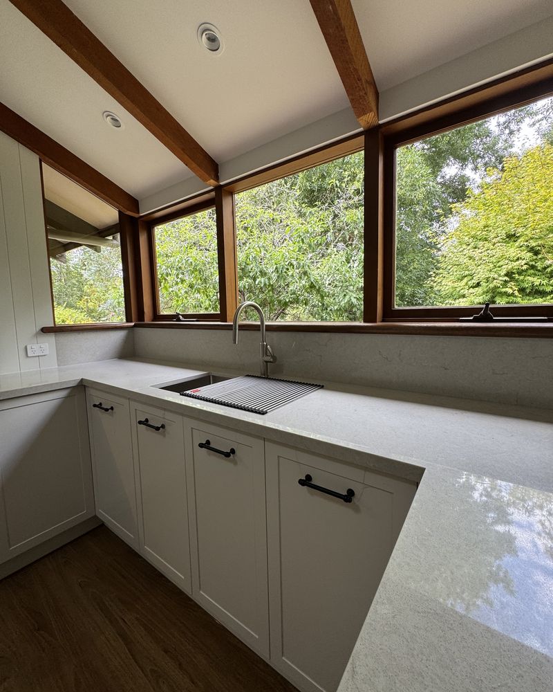 Corner of timber-framed windows above the sink and pale benchtop, with garden greenery outside