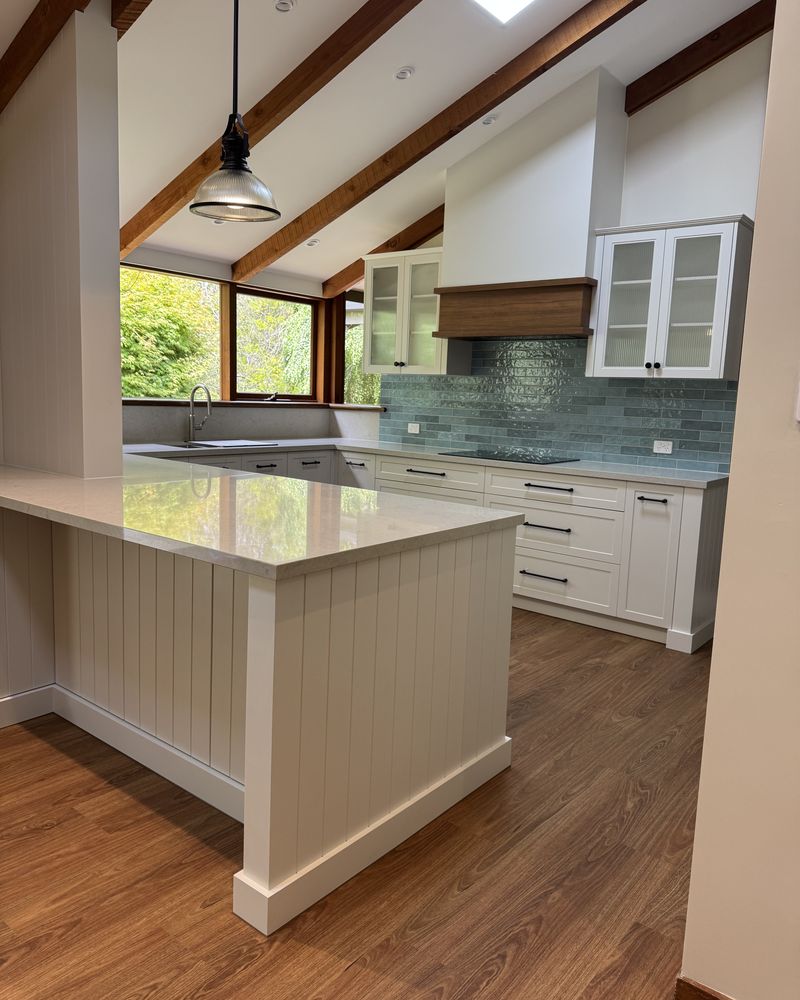 Island with a pale stone-look benchtop beneath a pendant light, with the timber rangehood, glazed uppers and blue-green tiled splashback beyond
