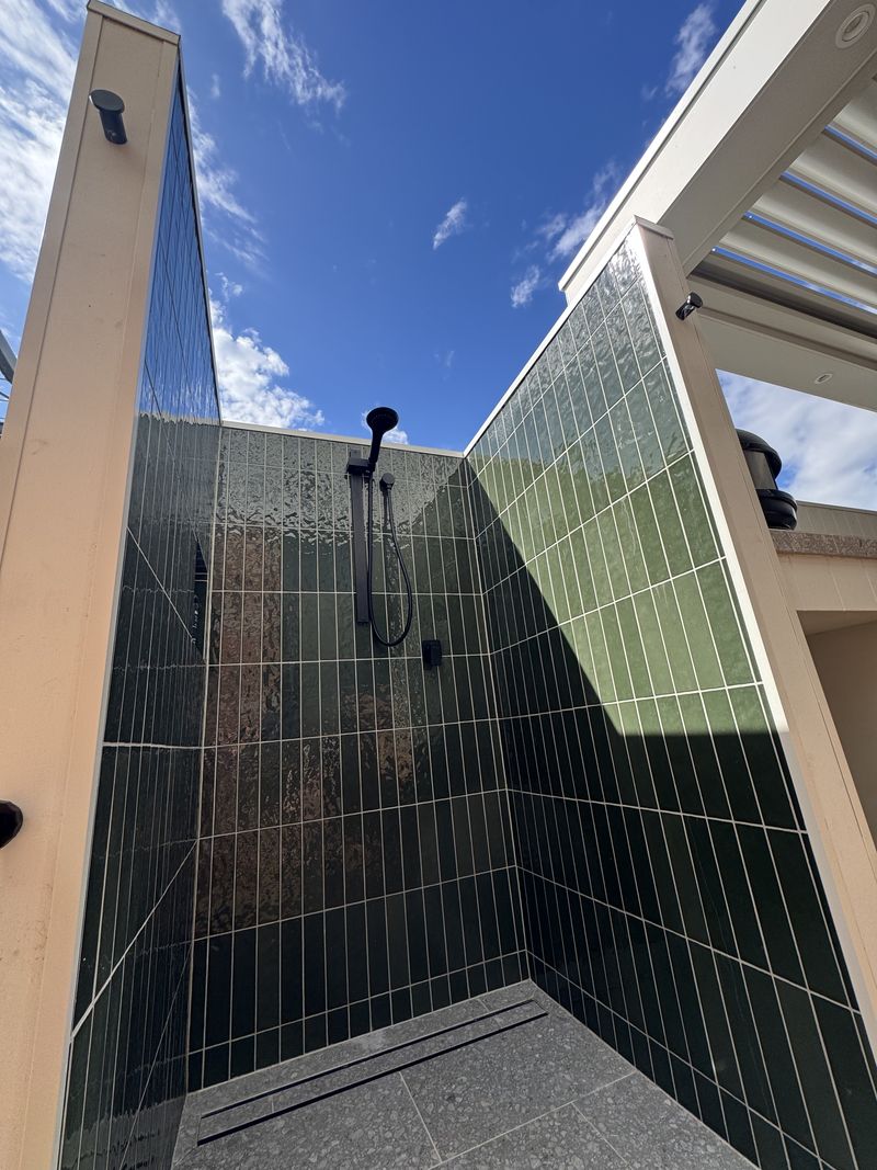 An open-air shower lined in glossy green vertical tile, with a matte black rail shower, a linear floor drain and blue sky overhead